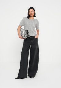 Γκρι πλεκτό πουλόβερ με κοντά μανίκια, συνδυασμένο με μαύρο παντελόνι φαρδιάς γραμμής. Κρατάει μια τσάντα φάκελο με υφή και μοτίβο φίδι σε μαύρο και άσπρο.