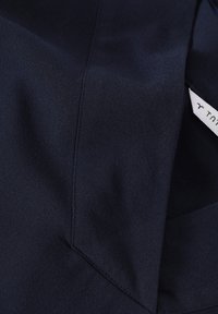 Tissu bleu marine avec une texture lisse, arborant un détail de couture et une petite étiquette avec le nom de la marque. Le matériau semble doux et tombe bien.