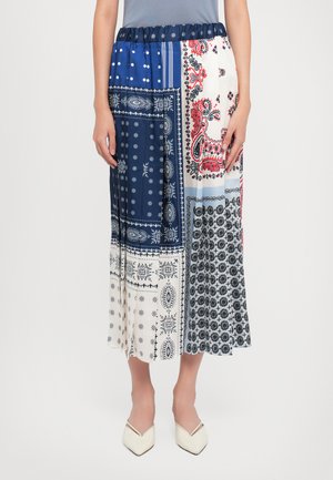 Frau in einem Patchwork-Midirock mit blauen, weißen, schwarzen und roten Paisley- und Blumendrucken, kombiniert mit weißen spitzen Schuhen.