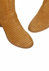 Jasnobrązowe zamszowe botki sięgające kostki z małymi otworami perforacyjnymi i niskim obcasem, pokazane z bliskiego kąta z góry na białym tle.