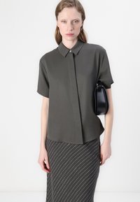 Camicia a maniche corte di colore verde oliva con colletto a bottoni, abbinata a una gonna a righe scure. La modella tiene una borsa rotonda nera.