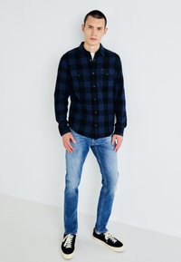 Cămașă în carouri albastre și negre din flanela, cu design cu nasturi, două buzunare la piept și mâneci lungi, purtată cu blugi de un albastru deschis și tenisi.