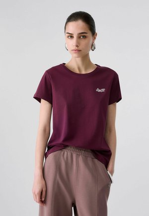 Giovane donna con capelli scuri legati in una coda di cavallo indossa una maglietta viola scuro e pantaloni marrone chiaro, grandi orecchini d'argento, in piedi davanti a uno sfondo grigio.