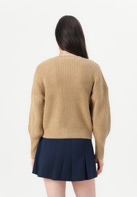 Beige neulepaita, jossa on rento leikkaus ja tiputetut olkapäät, yhdistetty tummansiniseen laskostettuun hameeseen. Teksturoitu pinta, rento muotoilu.