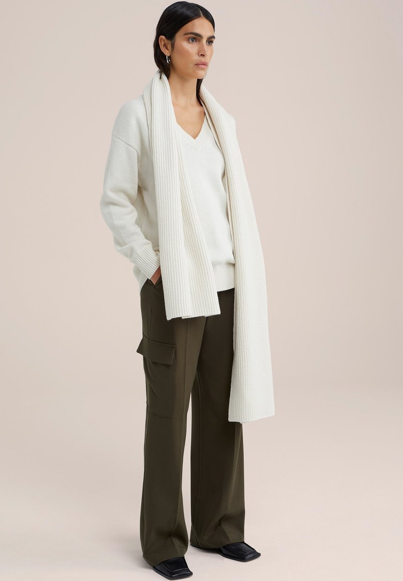 Strickpullover in Creme mit V-Ausschnitt, kombiniert mit olivgrünen, weit geschnittenen Hosen mit Seitentaschen und schwarzem Schuhwerk. Langer gerippter Schal drapiert.