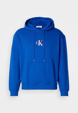 Blauwe pullover hoodie met een voorzak, verstelbare trekkoorden en het Calvin Klein-logo in het wit en rood, gecentreerd op de borst.