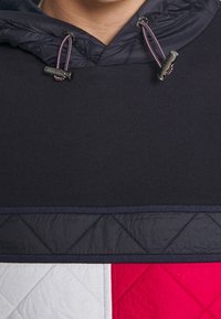 Zbliżenie na pikowaną kurtkę z czarnymi, białymi i czerwonymi panelami oraz ciemnonawigusiem z fioletowymi i białymi sznurkami ściągającymi z metalowymi końcówkami.
