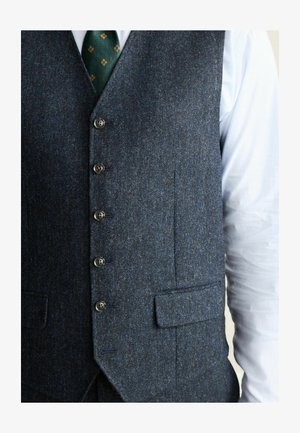 Tumedate sinise villase vest, millel on tekstuuriga viimistlus, viie nupuga ja kahe nurga all taskuga. Kande peal valge särk.