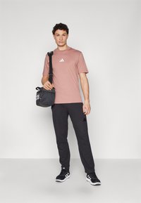 Herenoutfit met een lichtroze katoenen t-shirt met een witte logo, zwarte trainingsbroek en zwarte sportschoenen. De tas is zwart met een wit logo.