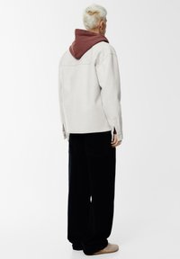 Lichtgrijze oversized jas met een bruine capuchon, gecombineerd met zwarte wijde ribfluwelen broek. Heeft een minimalistisch ontwerp en subtiele texturen.