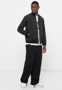 Black bomber jacket with ribbed cuffs and collar, layered over a white graphic t-shirt, paired with black cargo trousers and light sneakers.