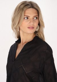 Schwarzes Shirt mit einem geometrischen Ausschnitt-Design, leichten Stoff und V-Ausschnitt, kombiniert mit strukturierten runden Ohrringen.