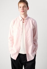 Ung mann iført en lys rosa skjorte med knapper over en hvit ribbestrikket tanktop og svarte bukser, stående med hendene i lommene mot en hvit bakgrunn.
