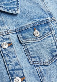 Ljusblå denimjacka med spetsig krage, två bröstfickor med silverfärgade tryckknappar och synliga sömmar över hela plaggdetaljer.