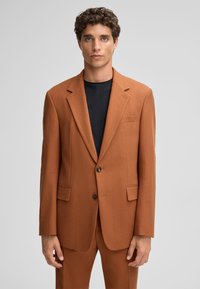 Rostfarbener Blazer mit strukturiertem Stoff, mit Revers, zwei vorderen Taschen und einer Zweiknopfverschluss. Getragen über einem schwarzen Hemd.