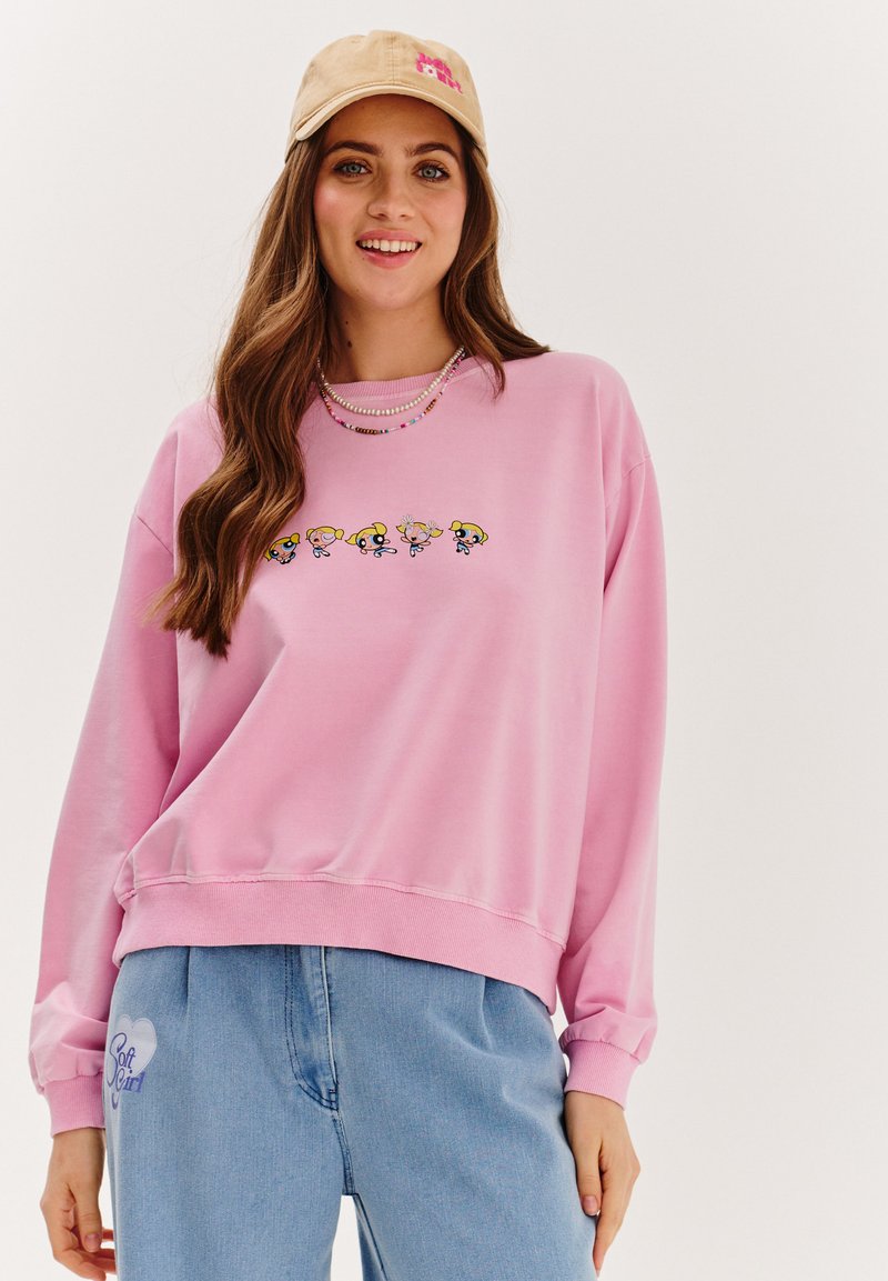 Růžová mikina se vyšitými karikaturními postavičkami přes hrudník. Kombinováno se světle modrými teplákovými kalhotami s podrobností logo srdce.