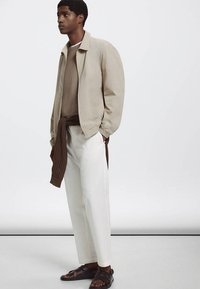 Homme debout portant une veste beige, un pull marron noué à la taille, un pantalon clair et des sandales marron foncé sur un fond clair uni.