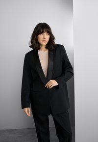 Čierne sako s lesklými saténovými klopami, jedným vreckom, kombinované s priesvitným topom s jemnými korálkovými ozdobami. Prispôsobený strih a štruktúrovaný dizajn.