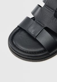 Zwarte leren sandaal met brede banden, een gladde textuur en een gevormd voetbed. Gestikt detail langs de randen, minimalistisch ontwerp.