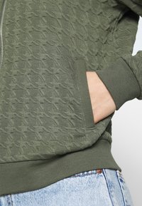 Olivgrön zip-up huvtröja med ett strukturerat mönster, ribbade cuffs och en sidoficka. Materialet verkar mjukt och något elastiskt.