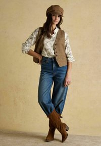 Brown plaid vest over a white shirt with bird print, paired with high-waisted blue denim and brown suede ankle boots.