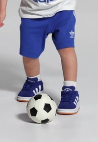 Shorts de sport bleus, t-shirt à logo blanc et bleu, baskets bleues avec des rayures blanches, se tenant à côté d'un ballon de foot noir et blanc.
