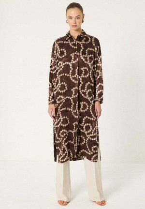 Žena v dlhej, tmavohnedej vzorovanej šnúrkovanej šaty cez biele nohavice so širokými nohavicami, stojaca bosá na jednoduchom bielom pozadí.