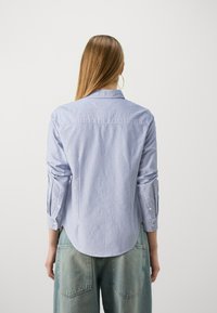 Camicia a righe blu e bianche con colletto a punta, maniche arrotolate e orlo curvo; abbinata a pantaloni larghi in denim chiaro.