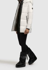 Chaqueta acolchada blanca con capucha, acentos negros y cremalleras, combinada con leggings negros y botas negras brillantes con cordones.