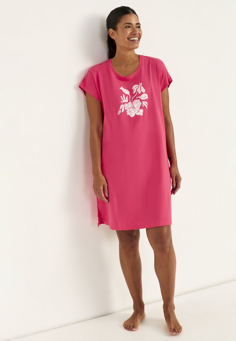 Lächelnde Frau mit dunklem Haar im Pferdeschwanz, die ein rosa Nachthemd mit Blumenmuster trägt und barfuß an einer weißen Wand steht.