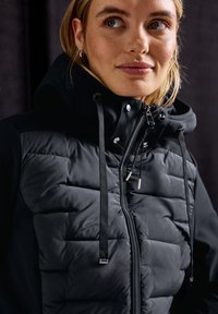 Schwarze gepolsterte Jacke mit Reißverschluss und Kordelzügen. Verfügt über eine Kapuze, glänzende Beschläge und ein strukturiertes Vorderteil. Glattes Material an den Seiten.