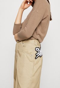 Beige, weit geschnittene Hose mit einem Patch auf der Gesäßtasche, der ein stilisiertes Schwarz-Weiß-Logo zeigt. Weicher, strukturiertes Gewebe.