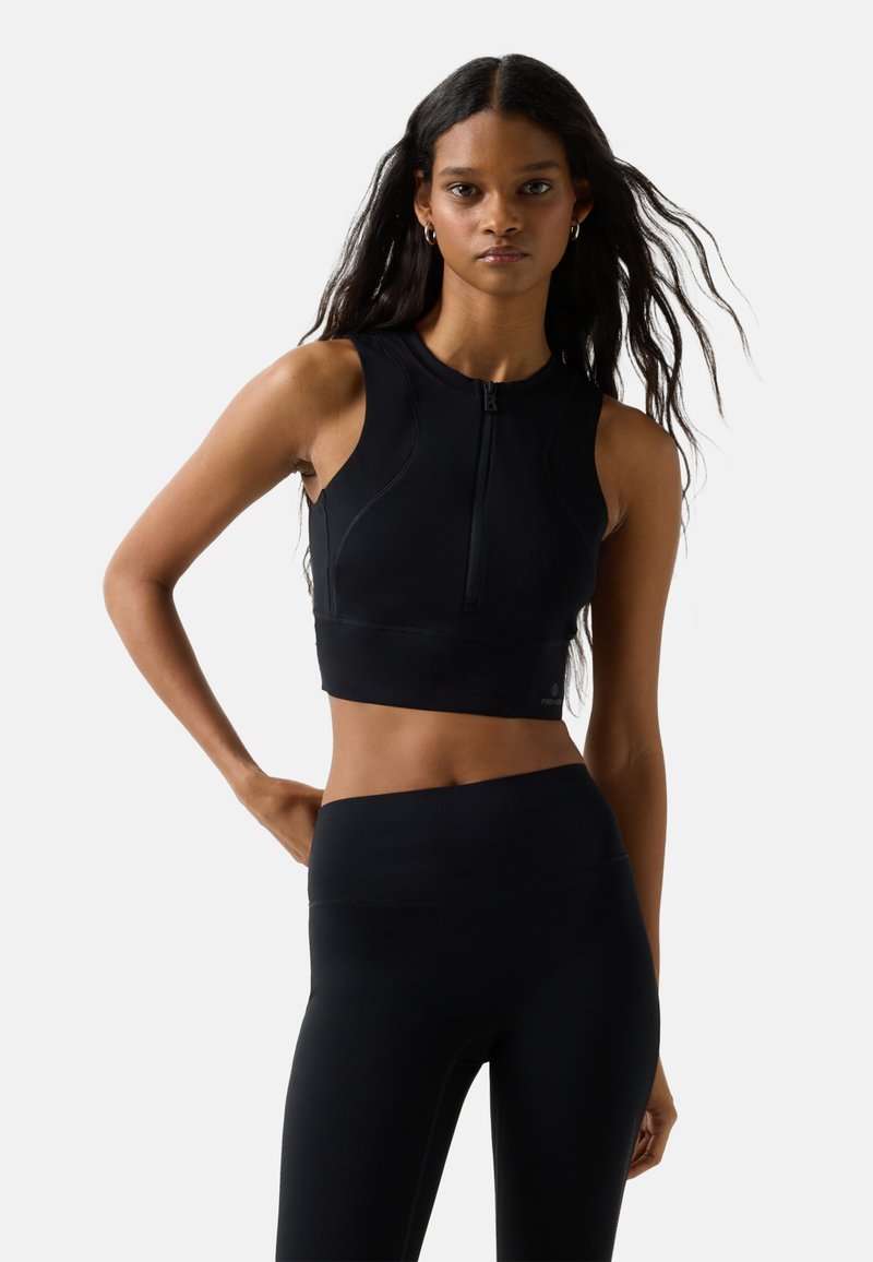 Frau mit langen dunklen Haaren, die ein schwarzes ärmelloses Crop-Top mit vorderem Reißverschluss und hoch taillierte schwarze Leggings trägt, steht vor einem weißen Hintergrund.