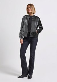 Czarna, błyszcząca kurtka bomberka z kołnierzem typu stójka, elastycznymi mankietami i przednimi kieszeniami, zestawiona z ciemnoniebieskimi dżinsami i czarnymi botkami za kostkę.