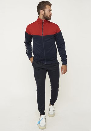 Uomo che cammina indossando una giacca con zip rossa e blu navy, pantaloni da jogging neri e sneakers bianche con un'etichetta blu sulla scarpa sinistra.
