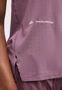 Κοντινό πλάνο σε μωβ διάτρητο αθλητικό τοπ με το λογότυπο H. Koumori φορεμένο από άτομο με σκουρόχρωμο δέρμα, που δείχνει λεπτομερείς ραφές και υφή υφάσματος.