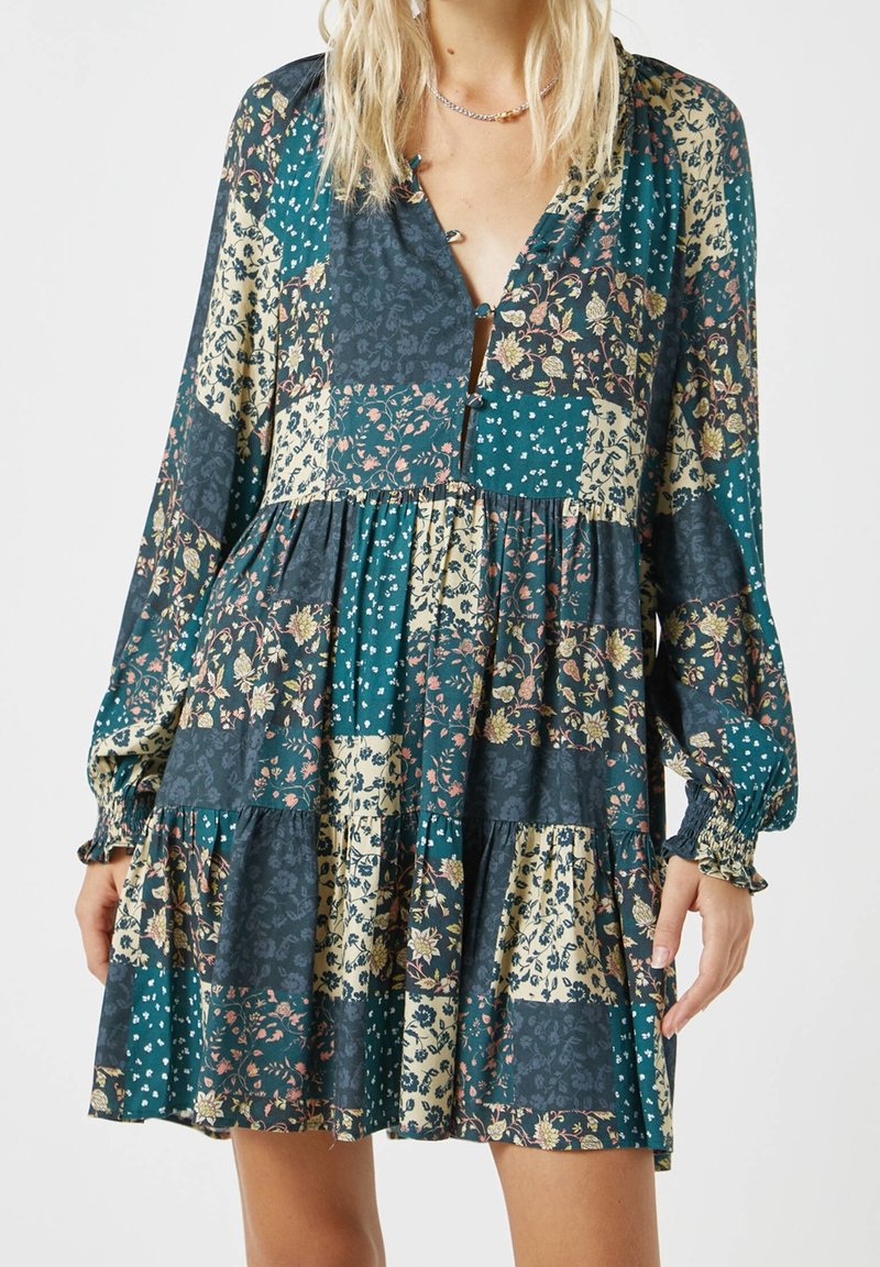 Robe à manches longues avec un décolleté en V, arborant un design en patchwork dans des motifs floraux et à pois teal, marine et beige. Jupe évasée et détail de boutons.
