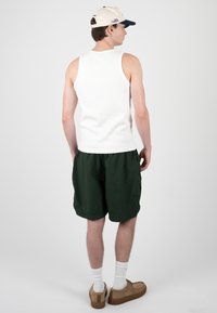 Jeune homme portant un débardeur blanc, un short vert foncé, des chaussures beiges, des chaussettes blanches et une casquette beige et noire, debout, dos à l’objectif, sur un fond uni.