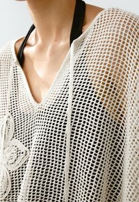 Zbliżenie na osobę w czarnym topie na ramiączkach pod beżowym swetrem z siateczki z kwiatowym haftem po lewej stronie.