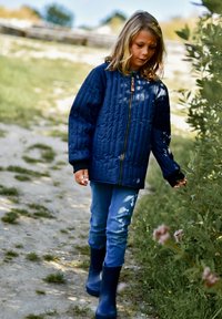 Tmavomodrá prešívaná bunda s predným zipsom a čiernymi manžetami, v kombinácii s modrými džínsami a tmavými gumákmi, nosená pri chôdzi po trávnatej ceste.