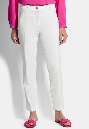 Frau trägt eng anliegende weiße Hose, leuchtend pinkes Langarmshirt und passende pinke geflochtene Slide-Sandalen, steht vor einem schlichten Hintergrund.