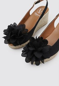 Sandalias de cuña negras tipo slingback con grandes acentos florales, plantilla de yute trenzado y forro interior suave. Parte trasera abierta para facilitar el uso.