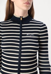 Navy and cream striped zip-up sweater with ribbed texture, high collar, and small embroidered "Tommy Jeans" logo on the chest.