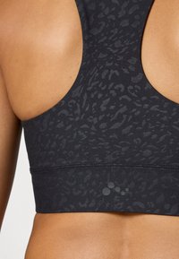 Zatvoren kadar gornjeg dijela leđa žene koja nosi crni sportski grudnjak s uzorkom i dizajnom racerback, s malim logom.