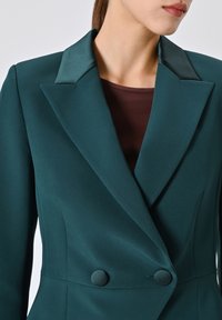 Blazer doppiopetto in verde acqua con risvolti in raso, bottoni arrotondati e design strutturato. Tessuto liscio con una texture sottile e vestibilità su misura.