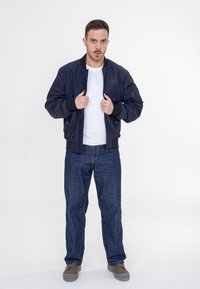 Marineblauer Bomberjacke mit Reißverschluss, gerippten Bündchen und Logo. Kombiniert mit einem weißen T-Shirt und dunklen Jeans, abgerundet mit grauen Schuhen.