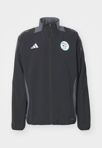Chaqueta deportiva negra con detalles grises, con cremallera completa, cuello alto, que presenta un logo redondo de fútbol y el distintivo de tres rayas en las mangas.