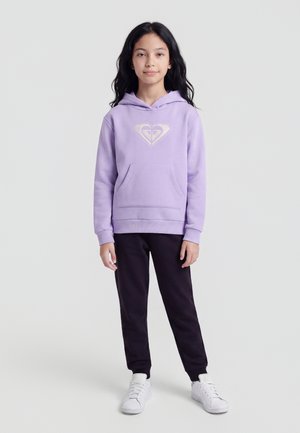 Fille aux longs cheveux foncés portant un sweat à capuche violet clair, un pantalon noir et des baskets blanches, debout devant un fond gris uni.