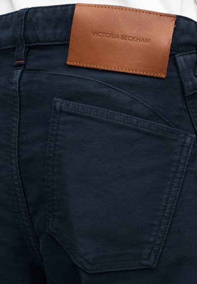Približen posnetek zadnjega žepa temno modrih hlač z rjavim usnjenim označevalcem, pritrjenim zgoraj, na katerem je vtisnjeno ime blagovne znamke "Victoria Beckham".
