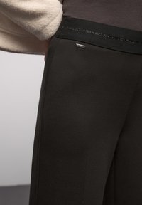 Schwarze, schmal zulaufende Hose mit glatter Textur und einem eng anliegenden Bund, der mit einem dünnen glitzernden Akzent verziert ist. Kleines Markenschild sichtbar.